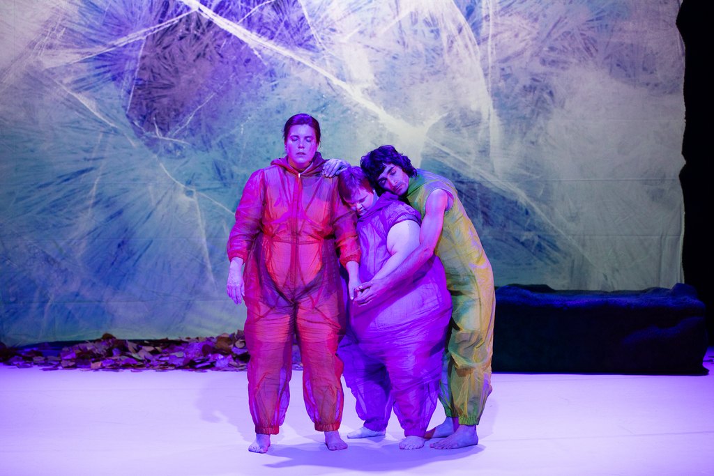 Tre personer i genomskinliga, färgskimrande dräkter står tätt samman på en scen, lutade mot varandra i ett stilla, sårbart ögonblick under blå-lila ljus.