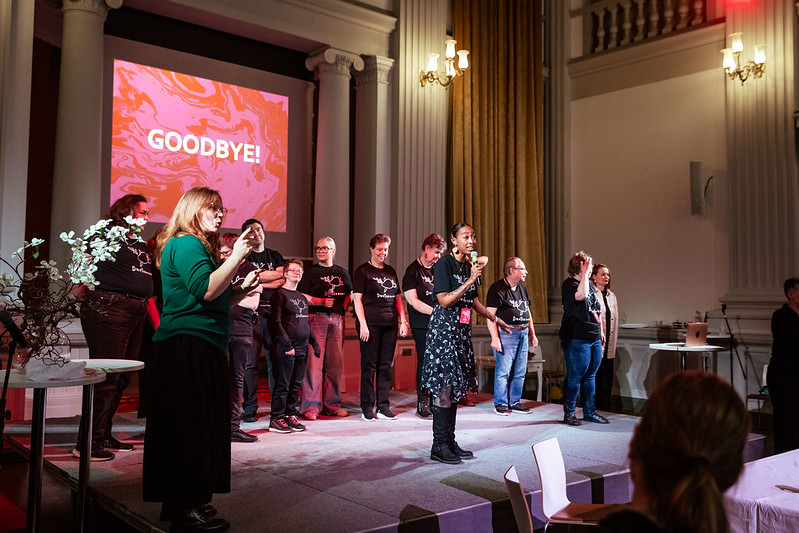 Group of performers on stage at a seminar, including a sign language interpreter on the far left, addressing the audience; behind them, a projected slide reads “GOODBYE!” in large letters.