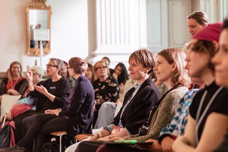Audience members seated at a seminar, listening attentively; one person signing while others watch and engage in a bright, formal indoor setting.