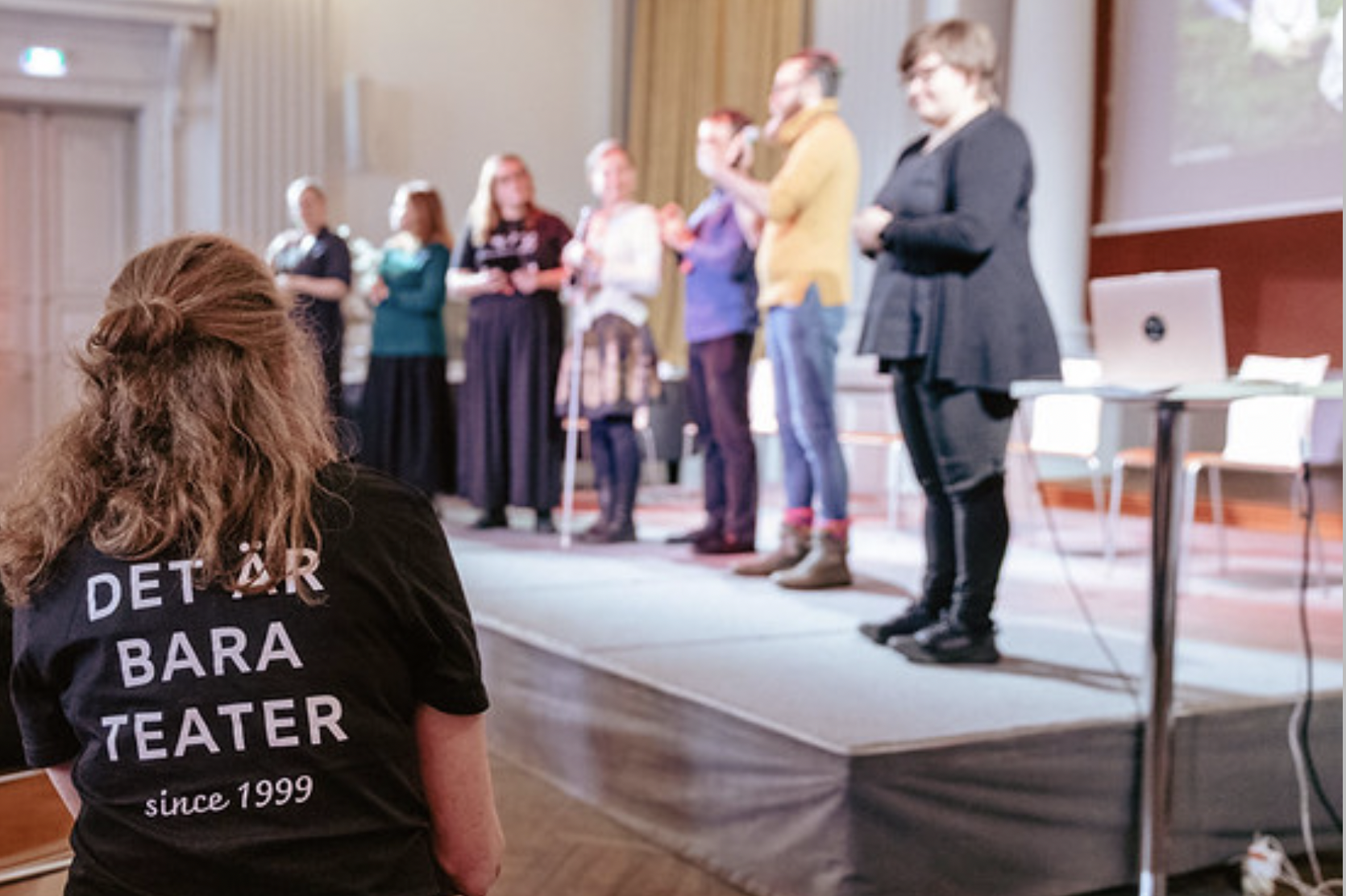 Audience member in the foreground wearing a T-shirt with the text “Det är bara teater” (“It’s only theatre”) on the back, watching a group of speakers on stage; several presenters stand in a row, some using sign language.