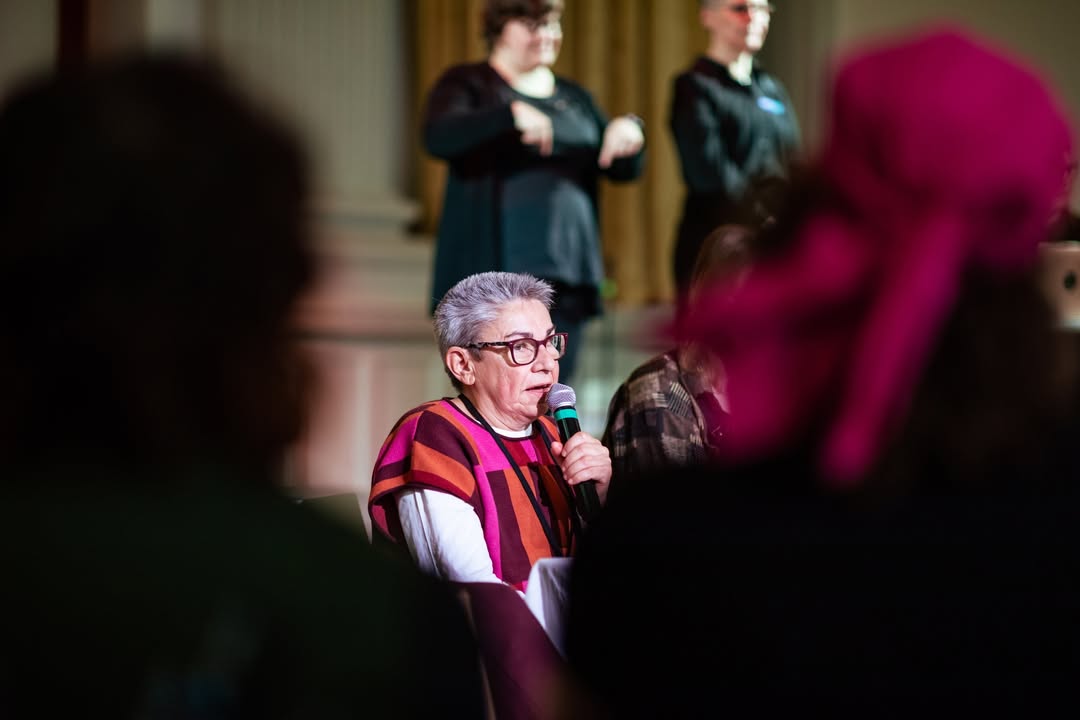 En föreläsningssal med höga vita pelare och varma gardiner är fylld av åhörare som sitter vända mot en scen. På scenen står tre personer: i mitten en talare som håller en mikrofon och papper, och på vardera sida två teckenspråkstolkar som gestikulerar tydligt. Bakom dem projiceras en stor röd presentationsbild med texten ‘Seminar on Creative accessibility in the Performing Arts, 20.10.2025’ samt organisatörernas logotyper. Stämningen är koncentrerad och professionell, och salen är upplyst av mjukt scenljus.